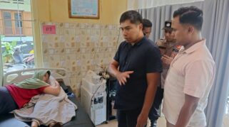 Gambar: Foto menunjukkan seorang pria sedang berbicara dengan seorang petugas polisi di sebuah ruangan rumah sakit. Terlihat seorang perempuan terbaring di tempat tidur di belakang mereka.
