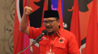 Seorang pria berbaju merah sedang berorasi dengan gestur semangat, sambil memegang mikrofon.
