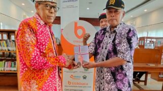 IGB Sudharma tersenyum memegang buku, mengenakan udeng merah putih, didampingi petugas perpustakaan.