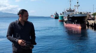 Seorang pria berdiri di pelabuhan dengan beberapa kapal kargo di latar belakang.